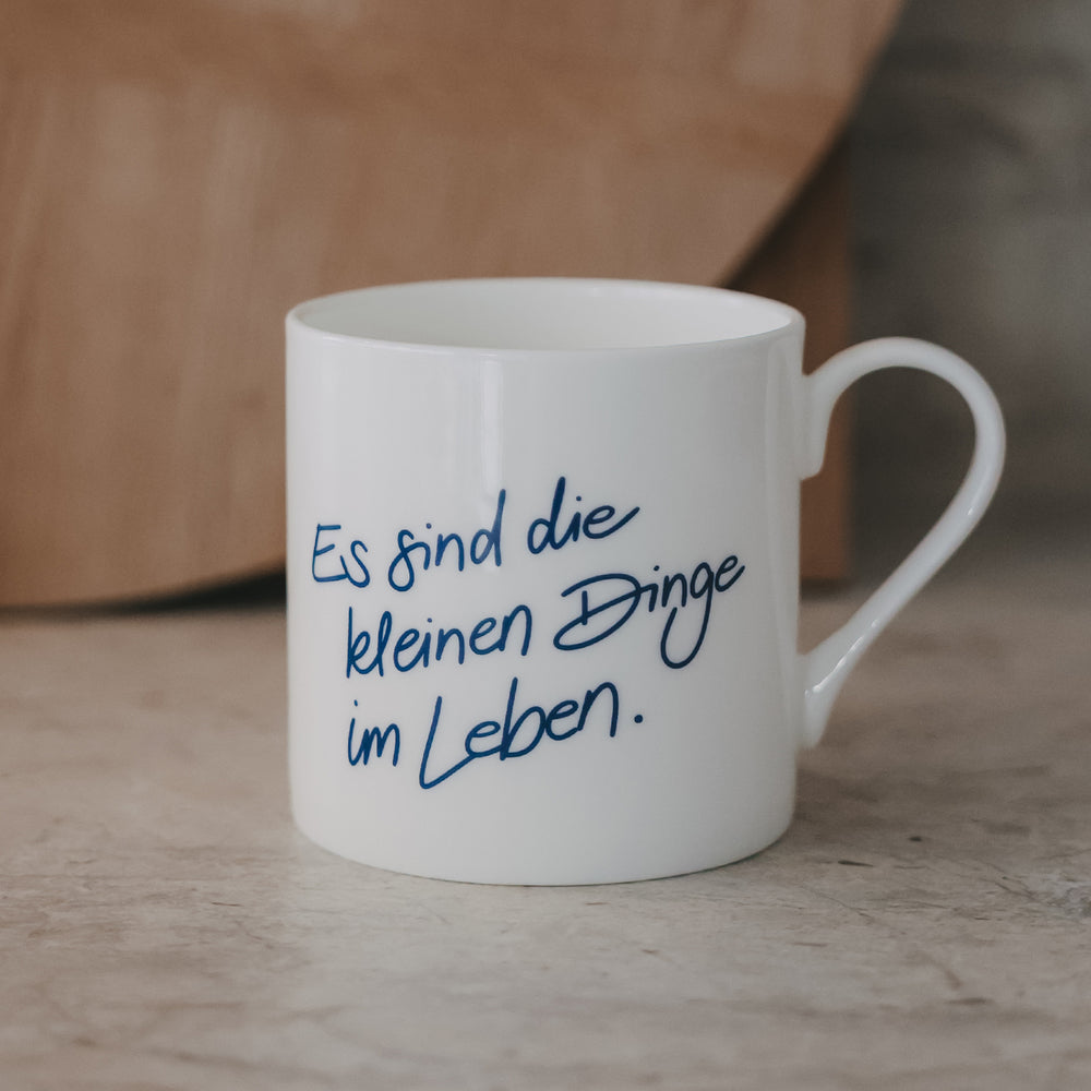 Weiße Tasse mit blauem Schriftzug „Es sind die kleinen Dinge im Leben“ vor warmem Holz-Hintergrund