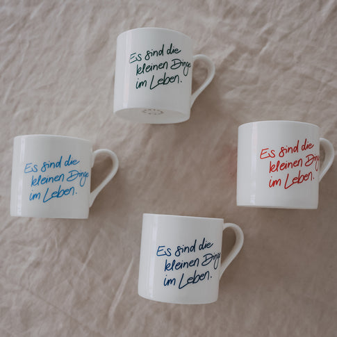 4 weiße Tassen mit Schriftzug „es sind die kleinen dinge im leben“ in verschiedenen Farben auf hellem Untergrund