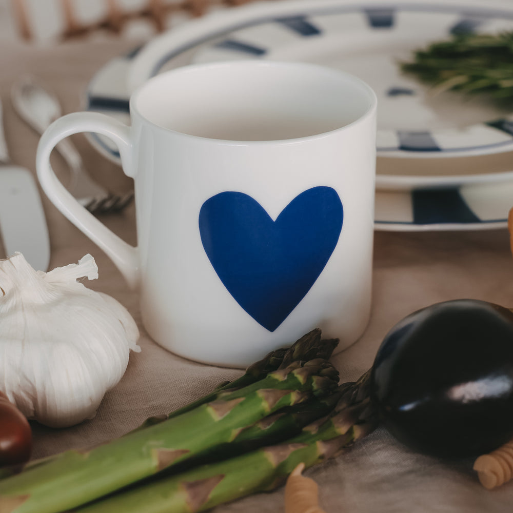 Weiße Tasse mit großem dunkelblauen Herz, dekoriert mit Spargel, Knoblauch und Geschirr im Hintergrund