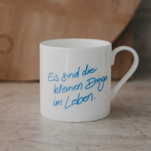 Weiße Tasse mit blauem Schriftzug „Es sind die kleinen Dinge im Leben“ vor warmem Holz-Hintergrund