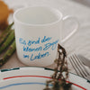 Weiße Tasse mit blauem Schriftzug „Es sind die kleinen Dinge im Leben“, dekoriert auf gedecktem Tisch mit Teller, Besteck und Spargel im Hintergrund