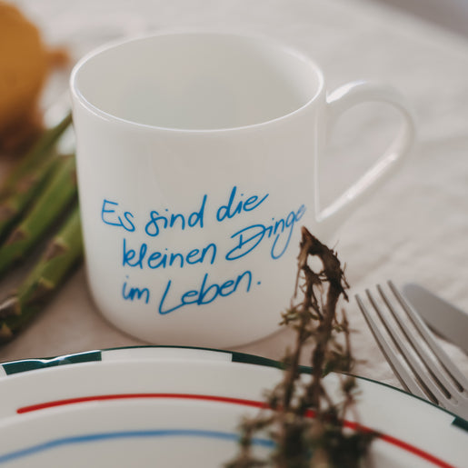 Weiße Tasse mit blauem Schriftzug „Es sind die kleinen Dinge im Leben“, dekoriert auf gedecktem Tisch mit Teller, Besteck und Spargel im Hintergrund