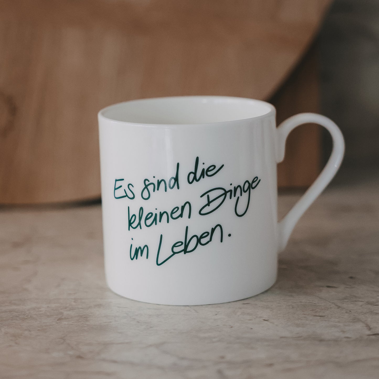 Weiße Tasse mit grünem Schriftzug „Es sind die kleinen Dinge im Leben“ vor warmem Holz-Hintergrund