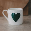 Weiße Tasse mit großem grünem Herzmotiv auf der Außenseite vor schlichtem Hintergrund