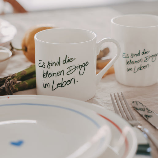 1 weiße Tasse und 1 weißer Becher mit grünem Schriftzug „Es sind die kleinen Dinge im Leben“ auf gedecktem Tisch mit Teller, Besteck und Spargel