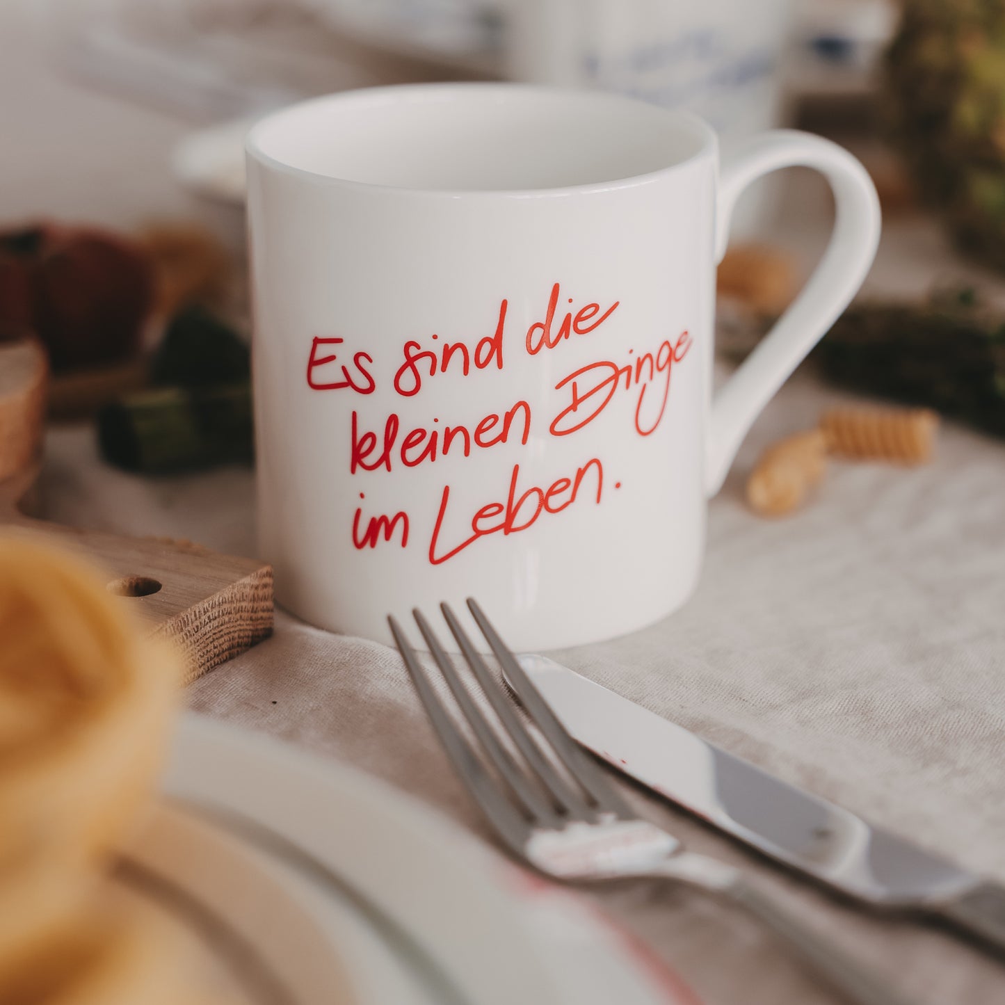 Weiße Tasse mit rotem Schriftzug „Es sind die kleinen Dinge im Leben“ auf gedecktem Tisch mit Besteck und Lebensmitteln
