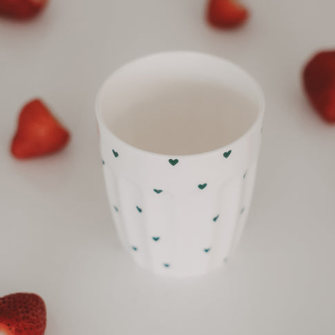 Keramikbecher mit grünen Herzen, umgeben von frischen Erdbeeren auf hellem Hintergrund
