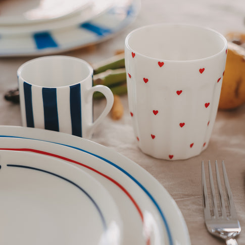 Herz-Becher neben gestreiftem Geschirr und Besteck, modernes Tischgedeck mit Keramikgeschirr