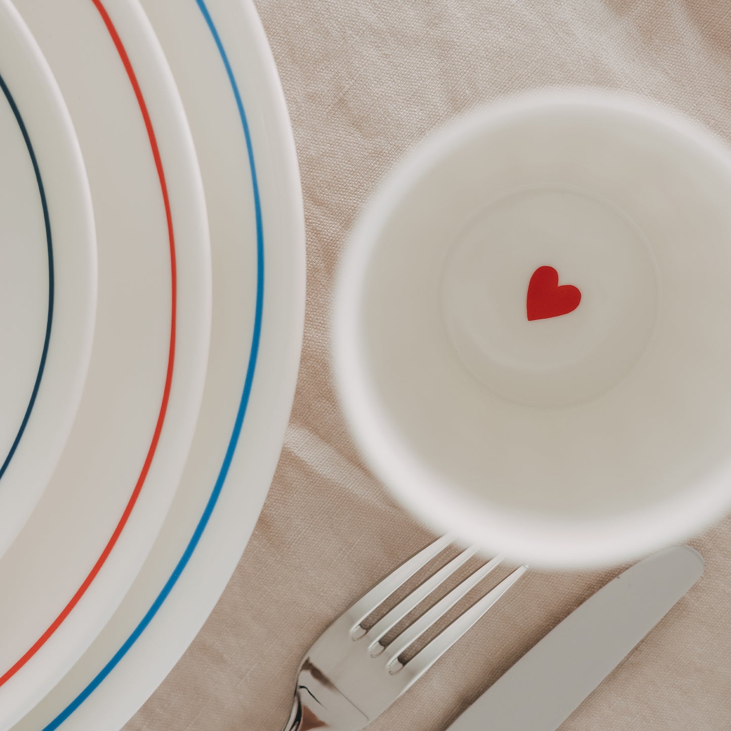 Detailaufnahme Becherboden mit rotem Herz, daneben Teller mit Streifen und Besteck auf Stoff