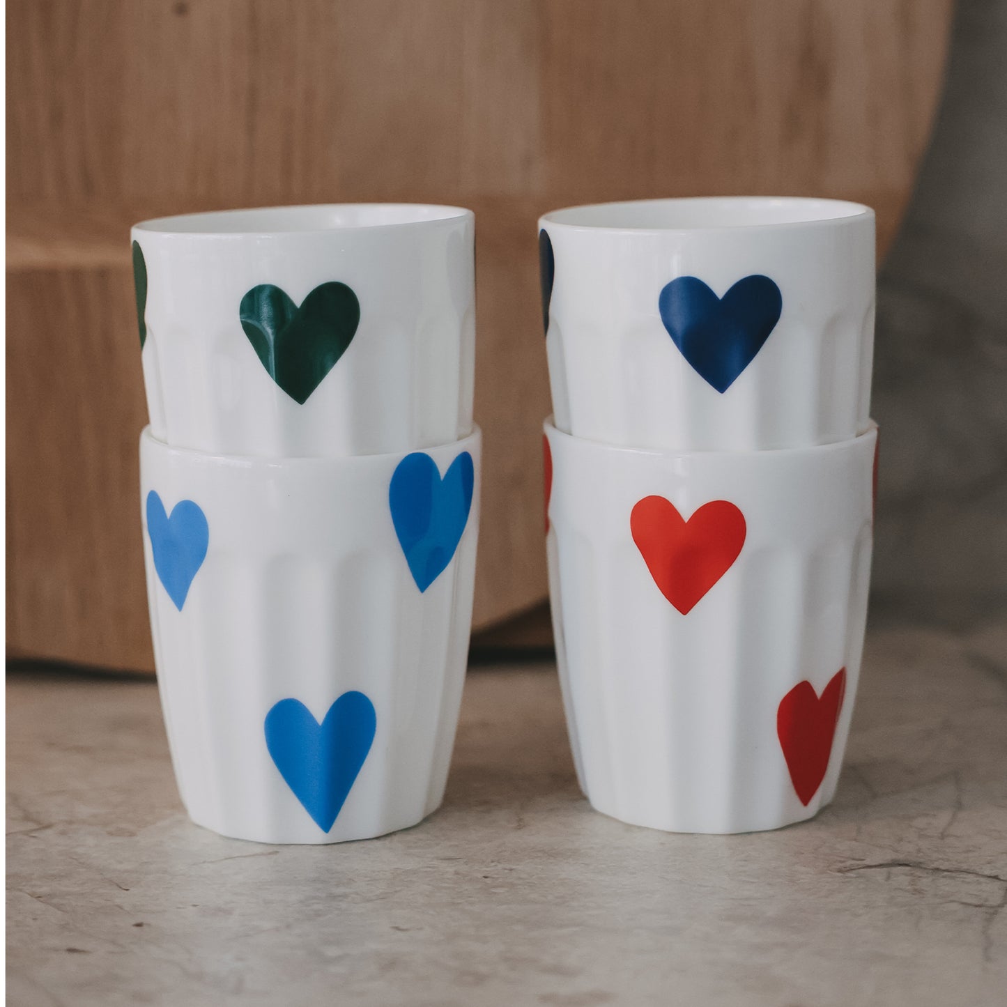 Stapelbare weiße Becher mit großen Herzmotiven in Blau, Grün und Rot auf Holzhintergrund
