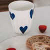 Weißer Becher mit blauem Herz neben Frühstück mit Brötchen und Erdbeere