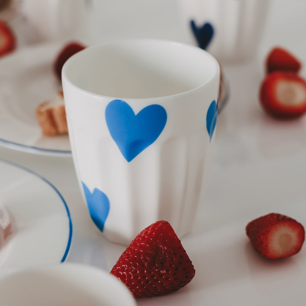 Keramikbecher mit blauem Herz neben Erdbeeren und Frühstück, stilvolle Tischszene