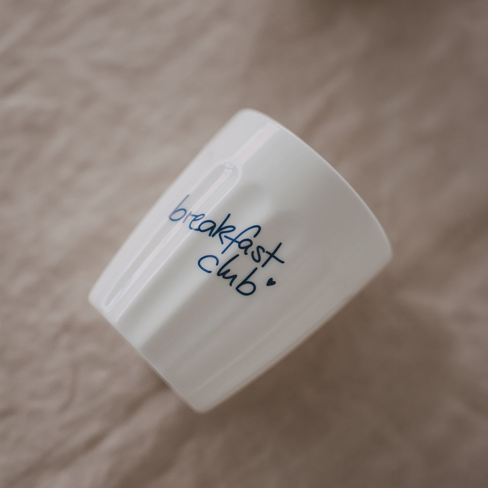 Weißer Keramikbecher „breakfast club“ mit blauem Schriftzug, schräg liegend auf hellem, weichem Stoffhintergrund
