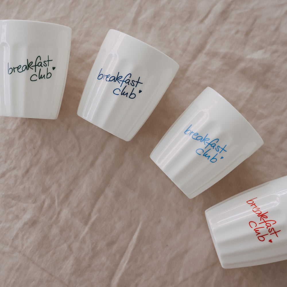 Keramikbecher „breakfast club“ in Blau und Grün dekorativ auf hellem Stoff platziert