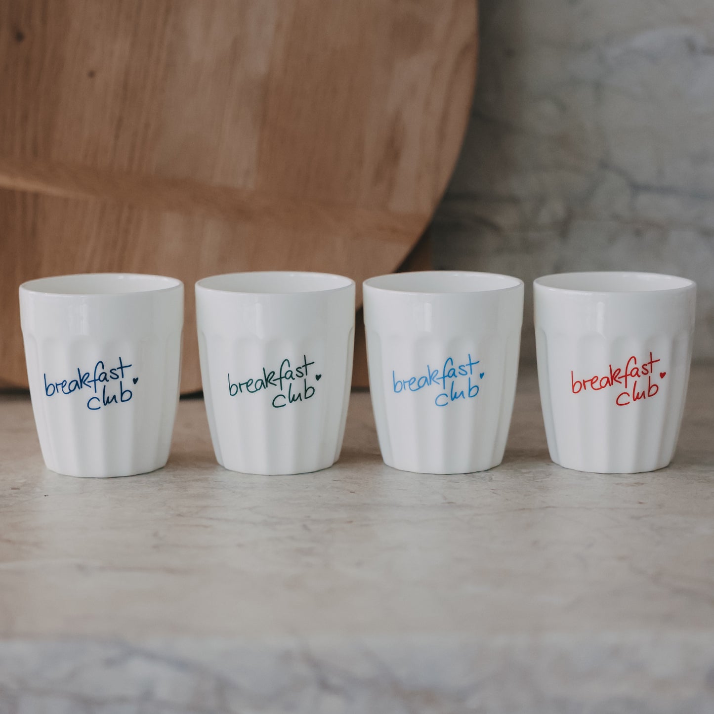 Vier weiße Keramikbecher „breakfast club“ mit farbigen Schriftzügen auf neutralem Hintergrund