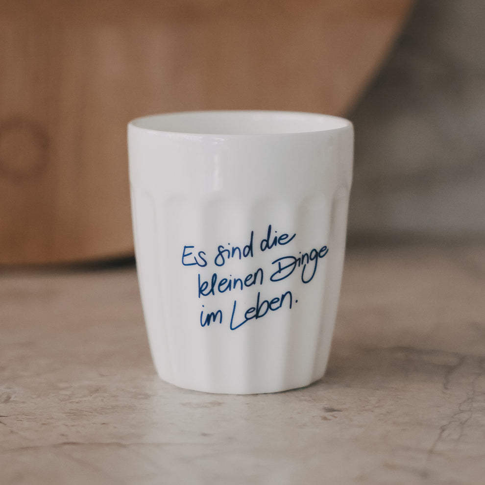 Weißer Keramikbecher mit Schriftzug „Es sind die kleinen Dinge im Leben“ in Blau auf hellem Untergrund