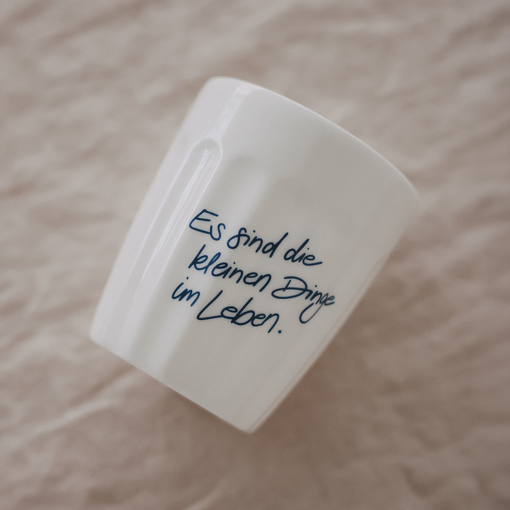 Weißer Becher mit Spruch „Es sind die kleinen Dinge im Leben“ in Blau, schräg liegend auf weichem Stoff