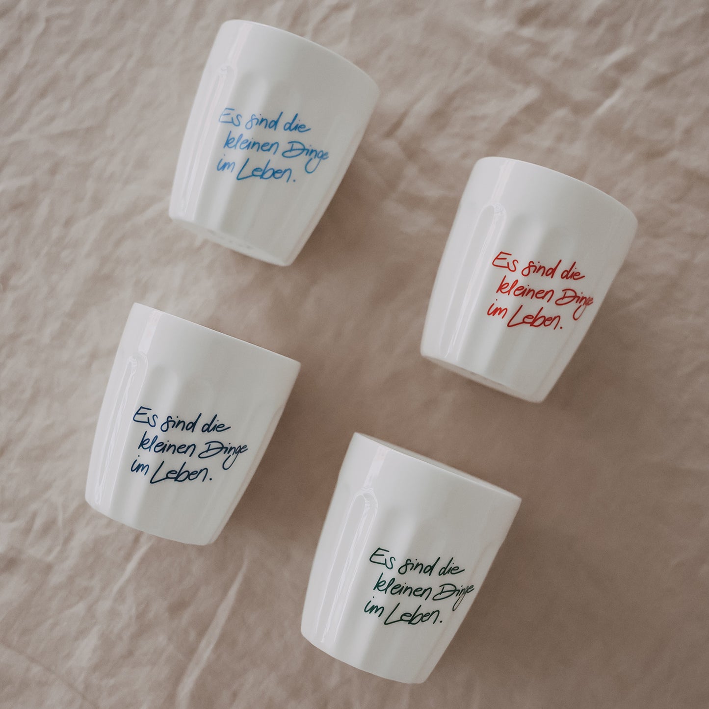 Vier weiße Keramikbecher mit farbigem Spruch „Es sind die kleinen Dinge im Leben“ dekorativ angeordnet