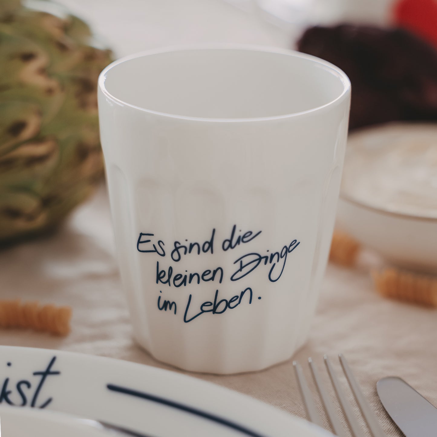 Weißer Keramikbecher mit Spruch „Es sind die kleinen Dinge im Leben“ in Blau, gedeckter Frühstückstisch im Hintergrund