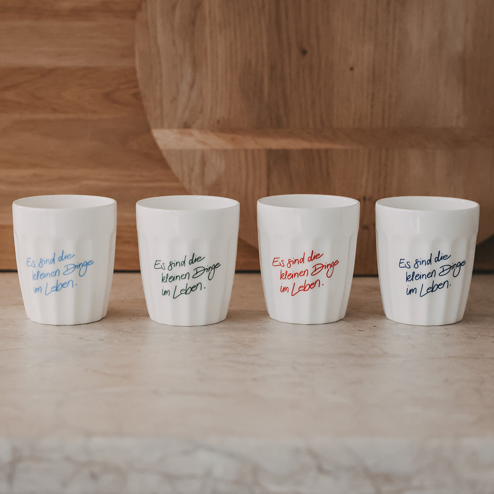Vier weiße Keramikbecher mit Spruch „Es sind die kleinen Dinge im Leben“ in verschiedenen Farben nebeneinander