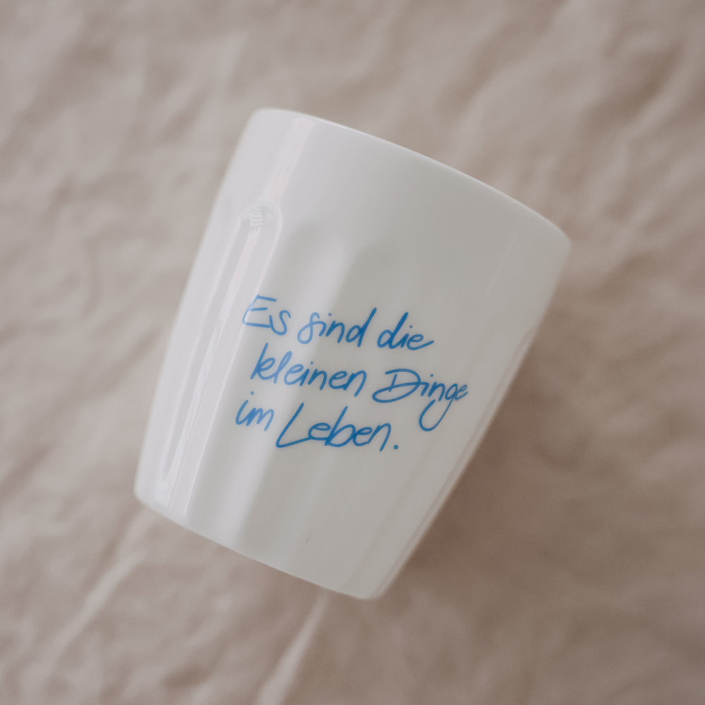 Weißer Keramikbecher mit Spruch „Es sind die kleinen Dinge im Leben“ in Hellblau, schräg liegend auf Stoff