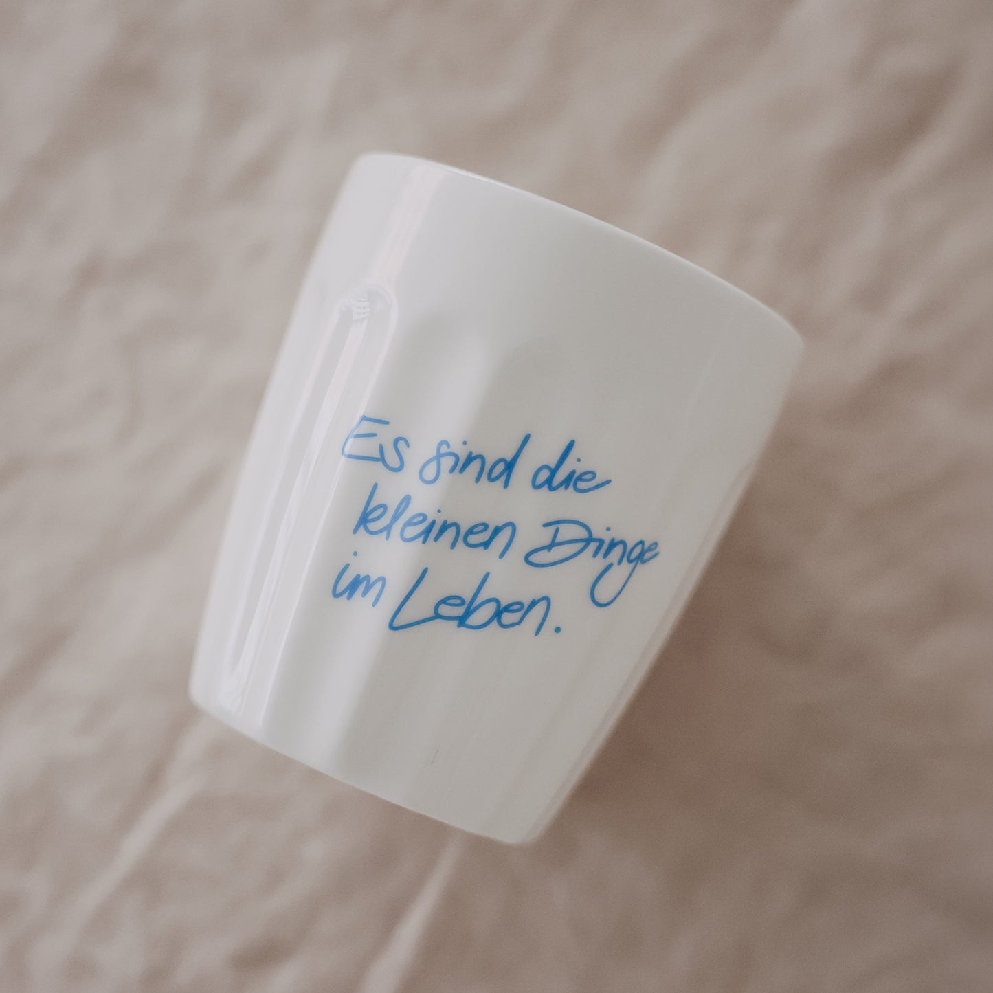 Weißer Keramikbecher mit Spruch „Es sind die kleinen Dinge im Leben“ in Hellblau, schräg liegend auf Stoff