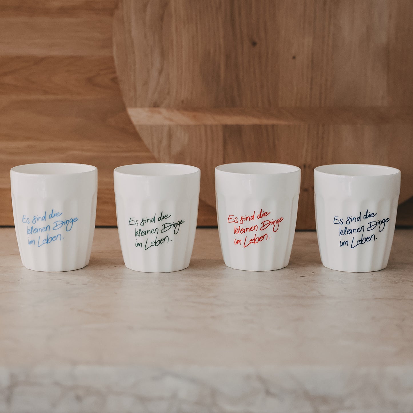 Vier weiße Becher mit Spruch „Es sind die kleinen Dinge im Leben“ in verschiedenen Farben nebeneinander