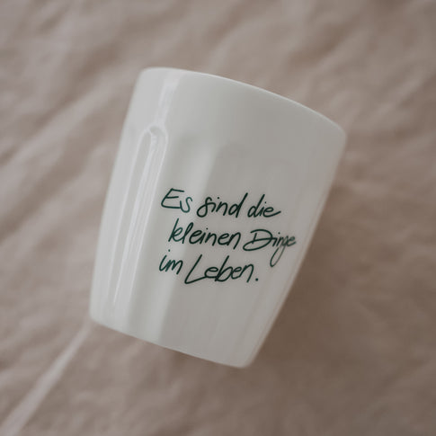 Weißer Keramikbecher mit grünem Schriftzug „Es sind die kleinen Dinge im Leben“, schräg auf Stoff platziert