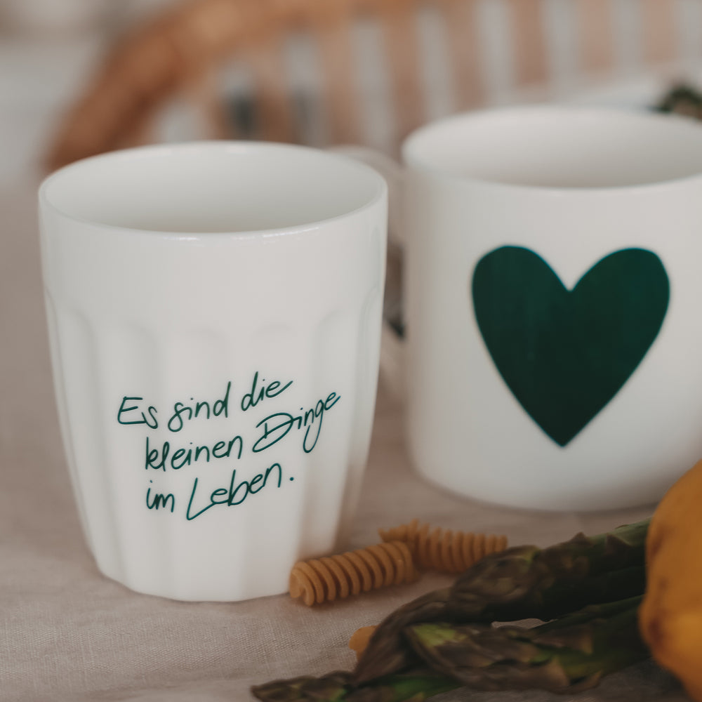 Zwei weiße Keramikbecher mit Spruch und grünem Herz, dekoriert mit Pasta und Gemüse auf hellem Stoff