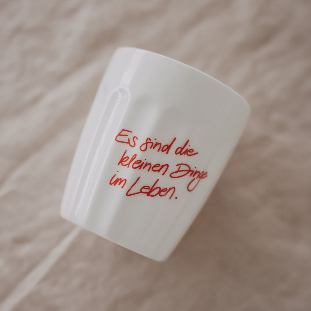 Weißer Keramikbecher mit rotem Schriftzug „Es sind die kleinen Dinge im Leben“, schräg auf Stoff platziert