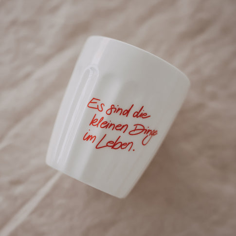 Weißer Keramikbecher mit rotem Schriftzug „Es sind die kleinen Dinge im Leben“, schräg auf Stoff platziert