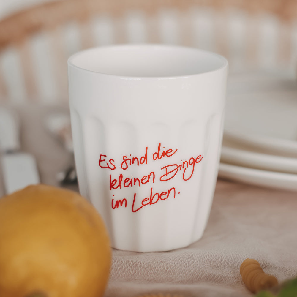 Weißer Keramikbecher mit rotem Spruch, dekoriert mit Gebäck und Pasta auf hellem Tischgedeck