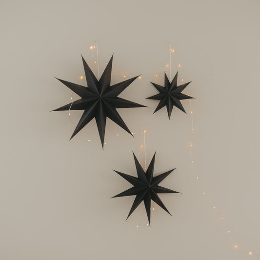 Drei schwarze Papiersterne in verschiedenen Größen mit warmer Lichterkette an heller Wand