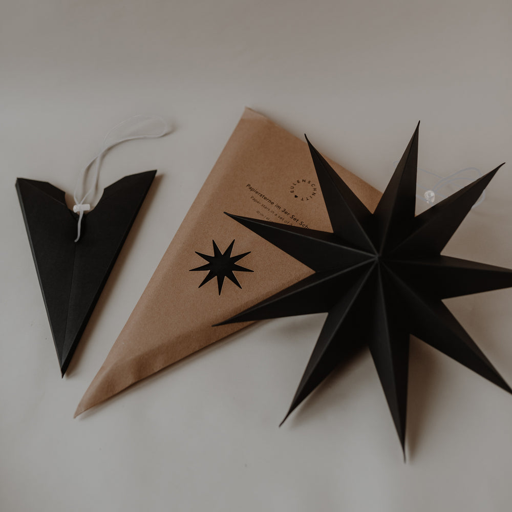 Schwarzer Papierstern mit Verpackung und zusammengefalteter Sternspitze mit weißer Aufhängung auf hellem Untergrund.