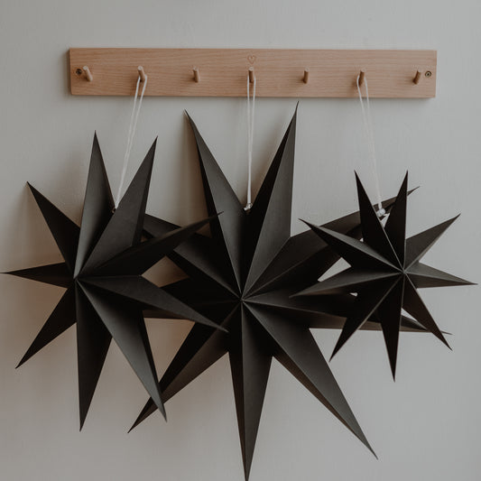 Drei schwarze Papiersterne hängen nebeneinander an einer Holzleiste mit kleinen Haken an einer weißen Wand.