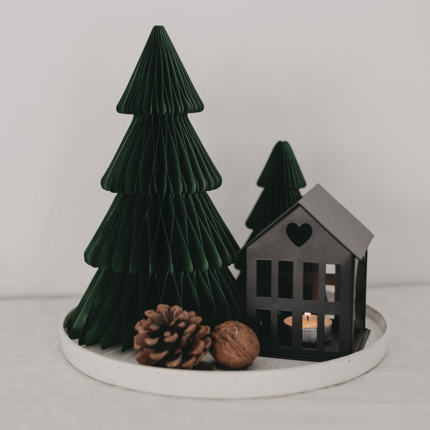 Dekoratives Weihnachtsarrangement auf einem runden weißen Tablett mit zwei dunkelgrünen Papiertannen, einem schwarzen Metallhäuschen mit Herz-Ausschnitt und einem brennenden Teelicht sowie einer Tannenzapfen- und Walnussdekoration.