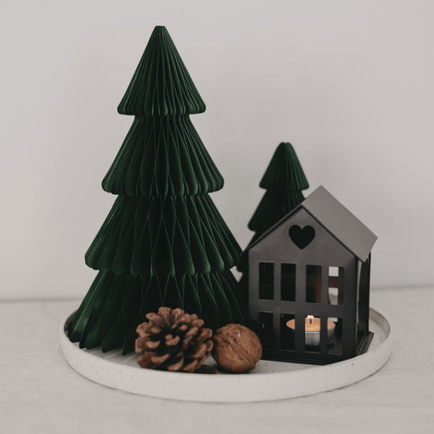 Dekoratives Weihnachtsarrangement auf einem runden weißen Tablett mit zwei dunkelgrünen Papiertannen, einem schwarzen Metallhäuschen mit Herz-Ausschnitt und einem brennenden Teelicht sowie einer Tannenzapfen- und Walnussdekoration.