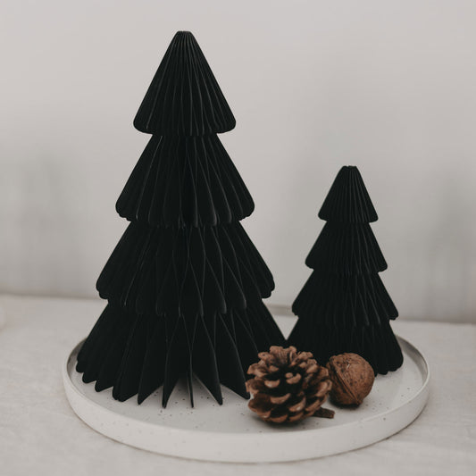 Dekoratives Weihnachtsarrangement mit zwei schwarzen Papiertannen in verschiedenen Größen auf einem weißen Steingutteller, ergänzt durch einen Tannenzapfen und eine Walnuss.