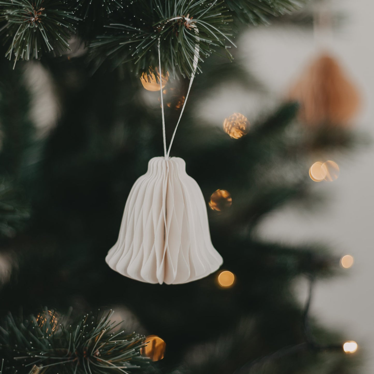 Weißer Papieranhänger in Glockenform mit Wabenstruktur am Tannenzweig mit Lichterkette