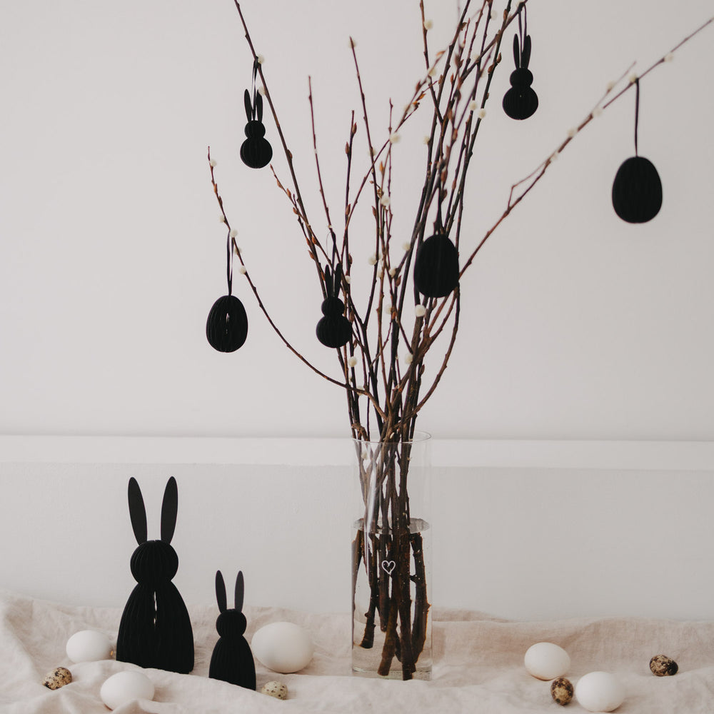 Schwarze Osteranhänger aus Papier an Zweigen im Glas mit Deko-Eiern auf hellem Stoff.