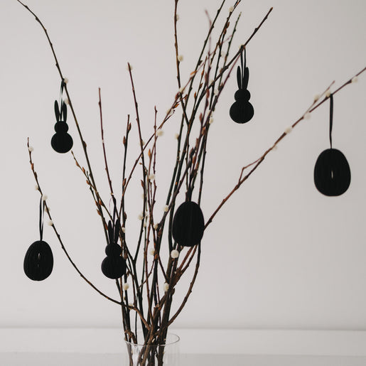 Schwarze Papier-Osteranhänger in Eiform und Hasenform an dekorierten Zweigen.