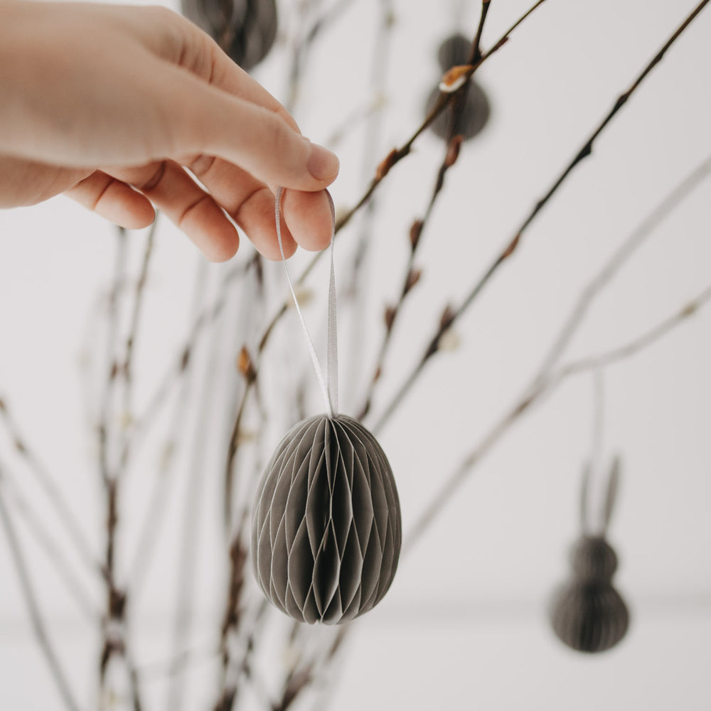 Hand hält grauen Papieranhänger in Eiform vor einem geschmückten Osterstrauch.