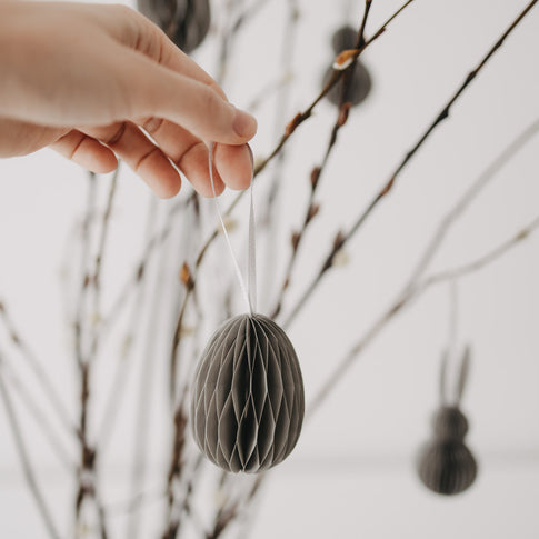 Hand hält grauen Papieranhänger in Eiform vor einem geschmückten Osterstrauch.
