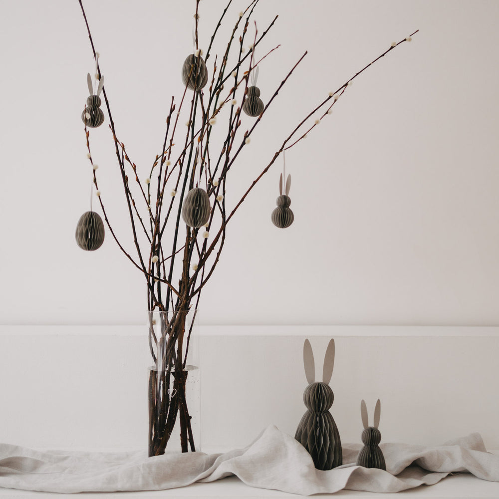 Grau­farbene Papierhasen vor Osterzweigen mit hängenden Anhängern als stilvolle Frühlingsdeko.