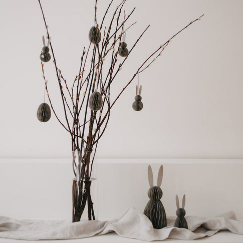 Grau­farbene Papierhasen vor Osterzweigen mit hängenden Anhängern als stilvolle Frühlingsdeko.