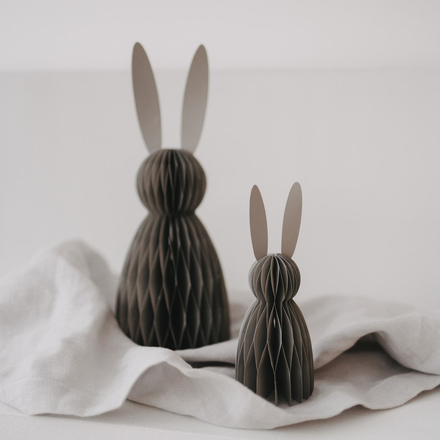 Zwei graue Papierhasen in kleiner und großer Größe als moderne Osterdeko auf hellem Stoff.