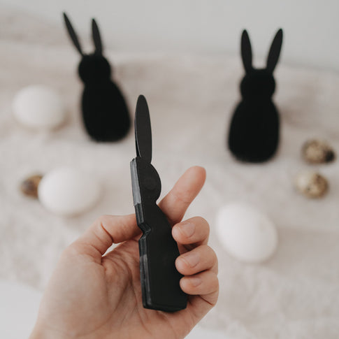 Schwarzer Papierhase in der Hand, weitere schwarze Papierhasen und Ostereier unscharf im Hintergrund.