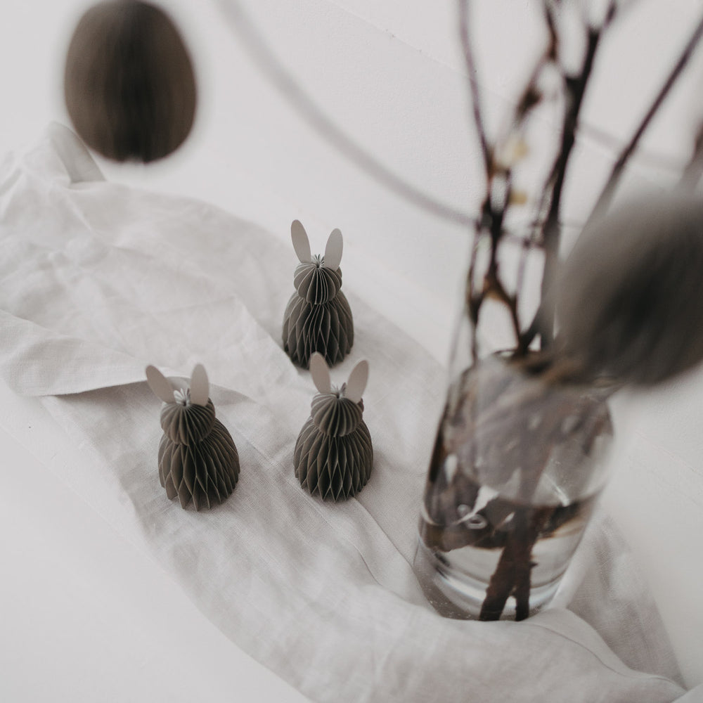 Graue Papierhasen und Osterdeko neben Ästen in Vase, stilvoll auf weißem Stoff dekoriert.