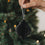 Schwarzer Papieranhänger in Kugelform wird vor einem festlich beleuchteten Weihnachtsbaum gehalten