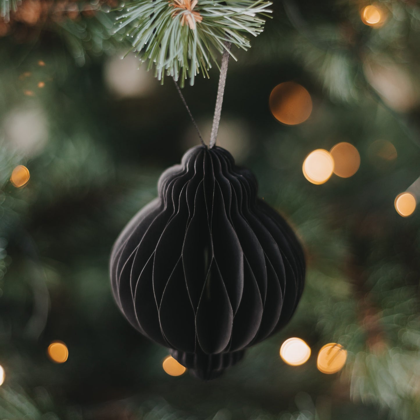 Schwarzer Papieranhänger in Kugelform am Weihnachtsbaum mit warm leuchtender Lichterkette